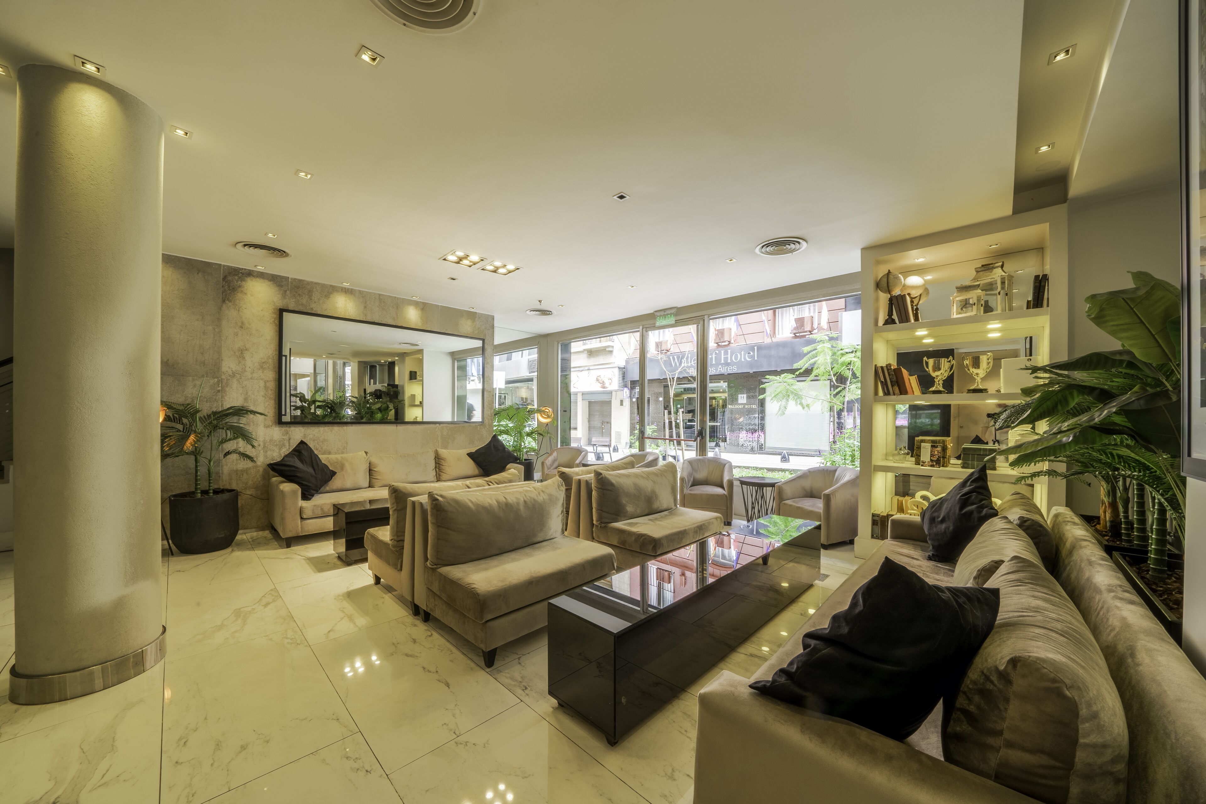 A modern hotel lobby with a marble floor, beige sofas, a coffee table, and a bookshelf with trophies.