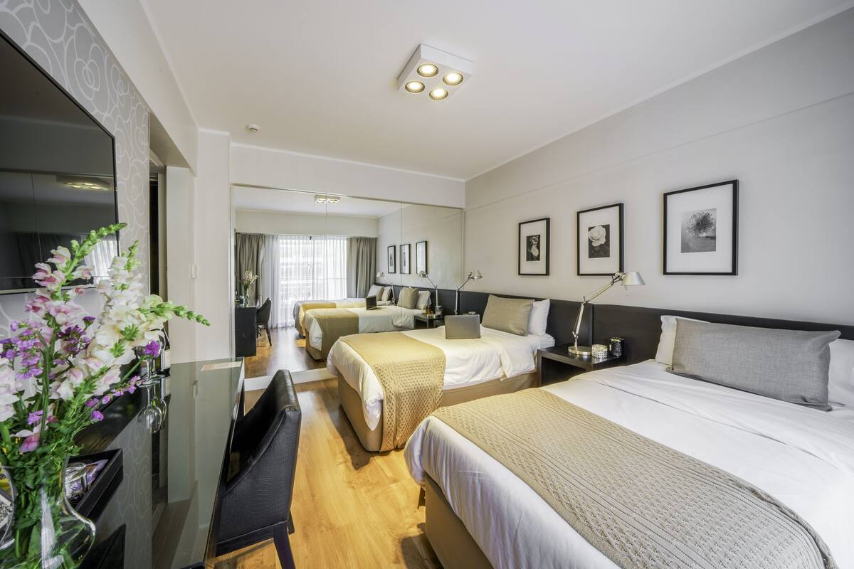 Habitación de hotel con una cama grande, una cama más pequeña, un escritorio y un televisor. Hay cuadros en la pared y un florero con flores en el escritorio.