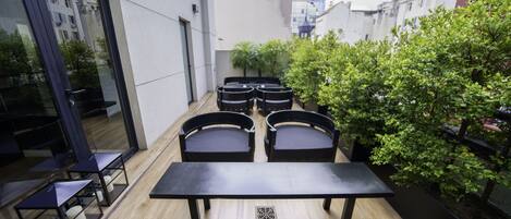Un patio con suelo de madera, una mesa en el centro y cuatro sillas. Hay plantas en macetas y se ve edificios al fondo.
