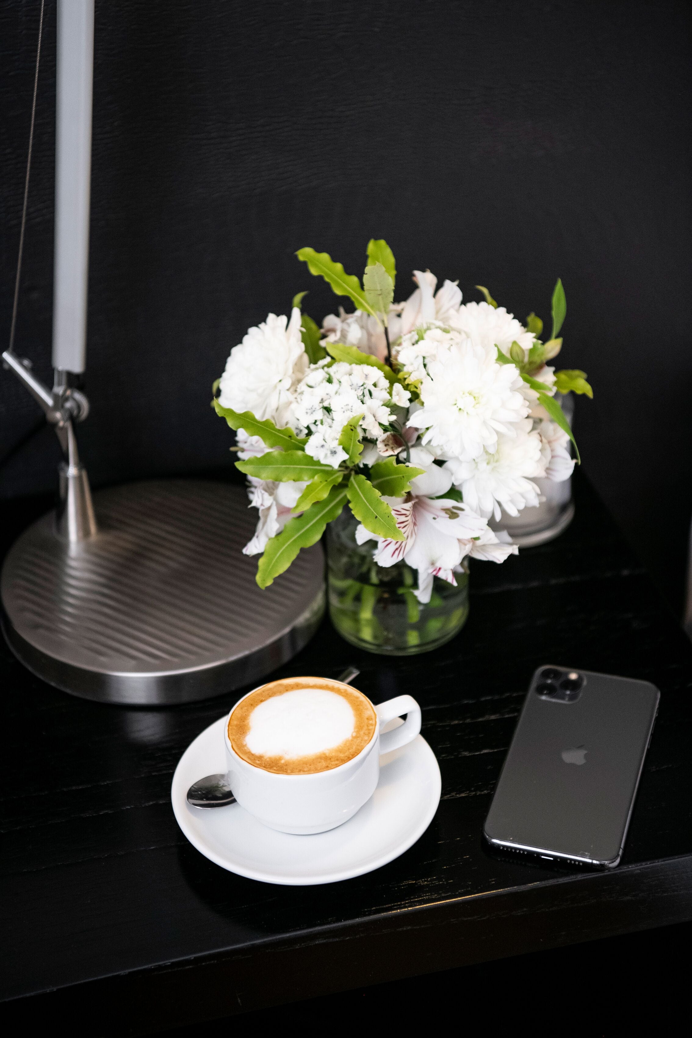 A cup of coffee with a latte art design, a smartphone, and a vase with white flowers on a black table.