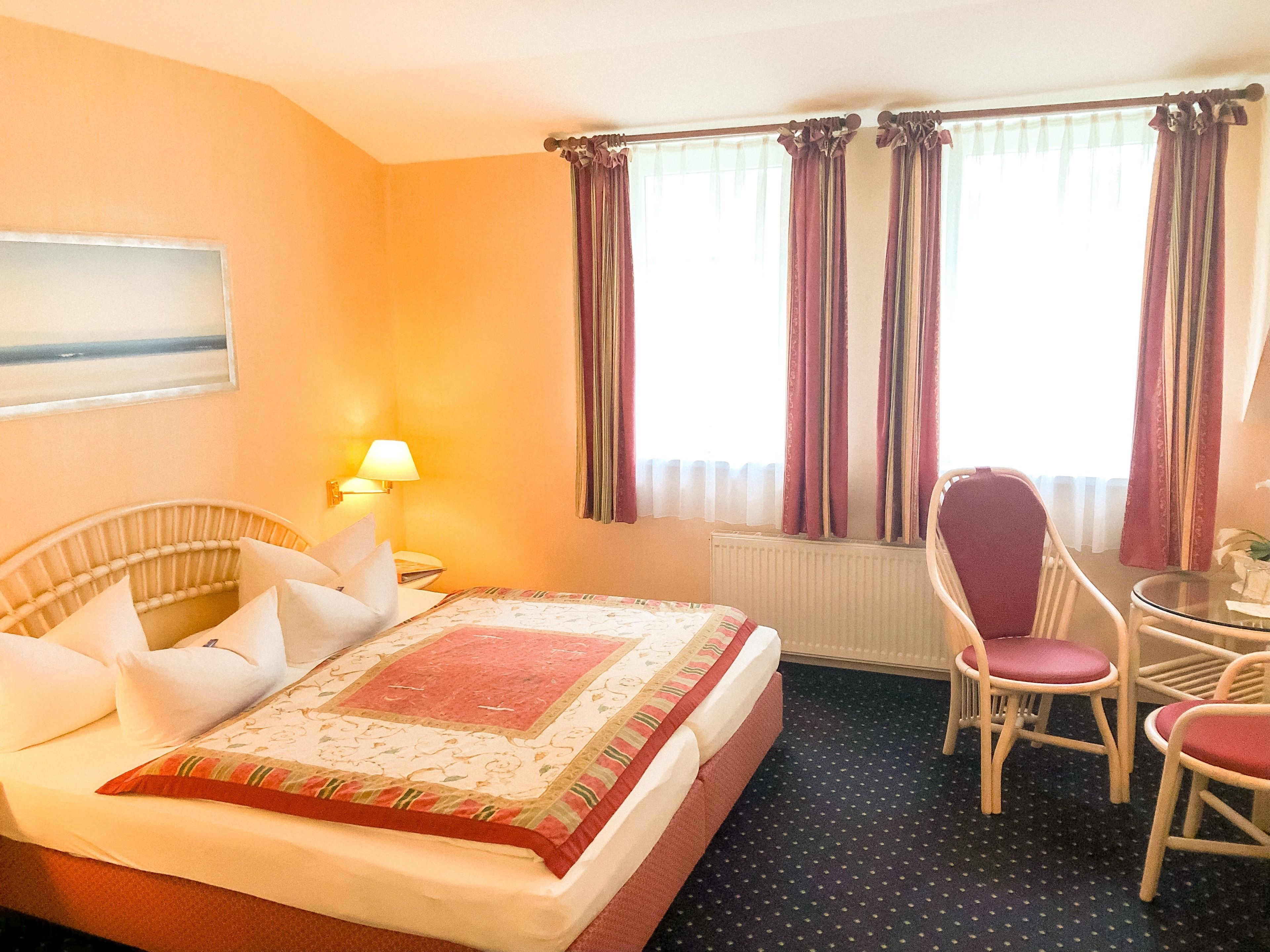 A hotel room with a bed, a desk, two chairs, a lamp, and a window with curtains.