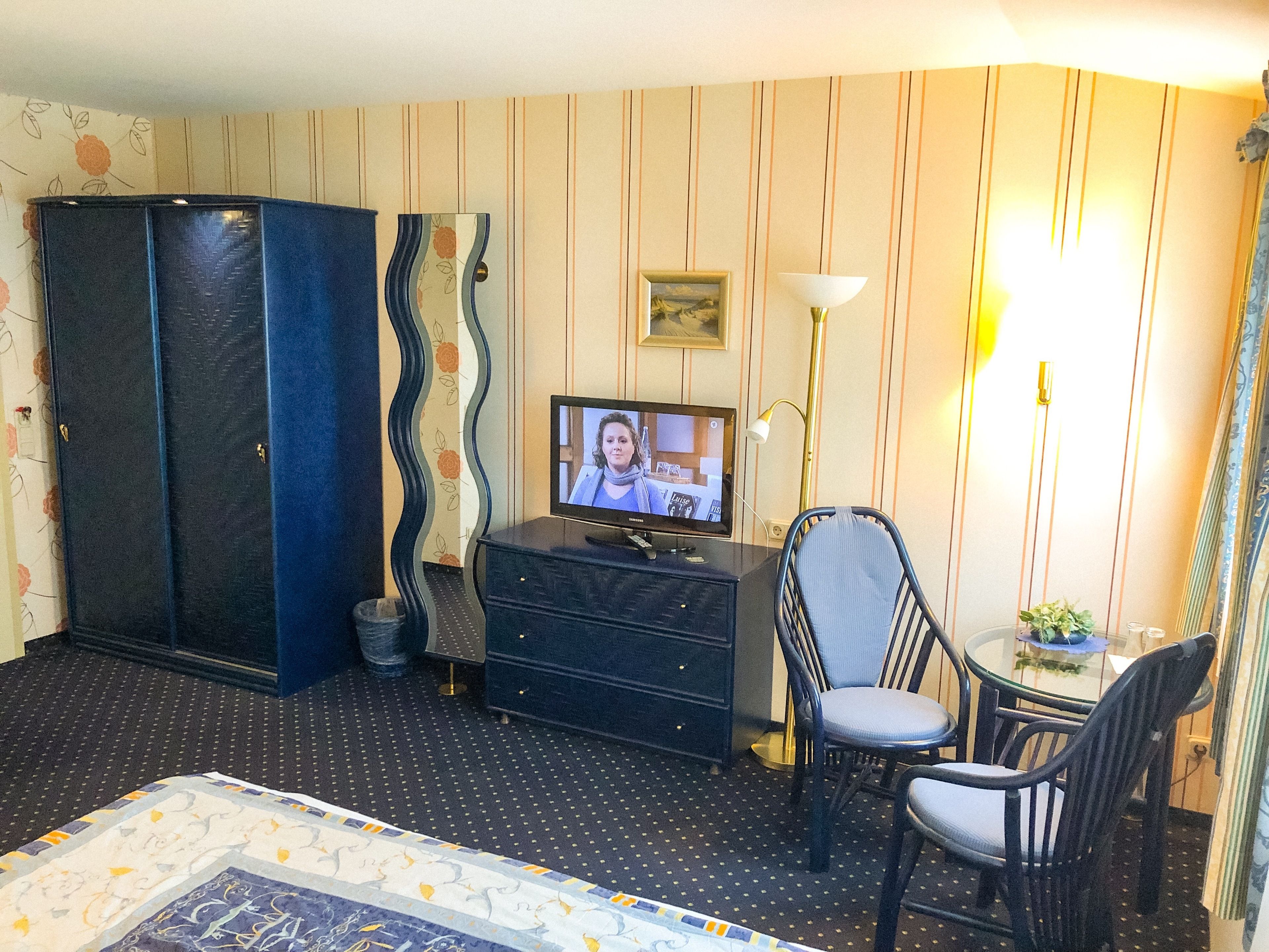 Hotellihuone, jossa on sänky, televisio, lipasto, tuoli, pieni pöytä ja lamppu.