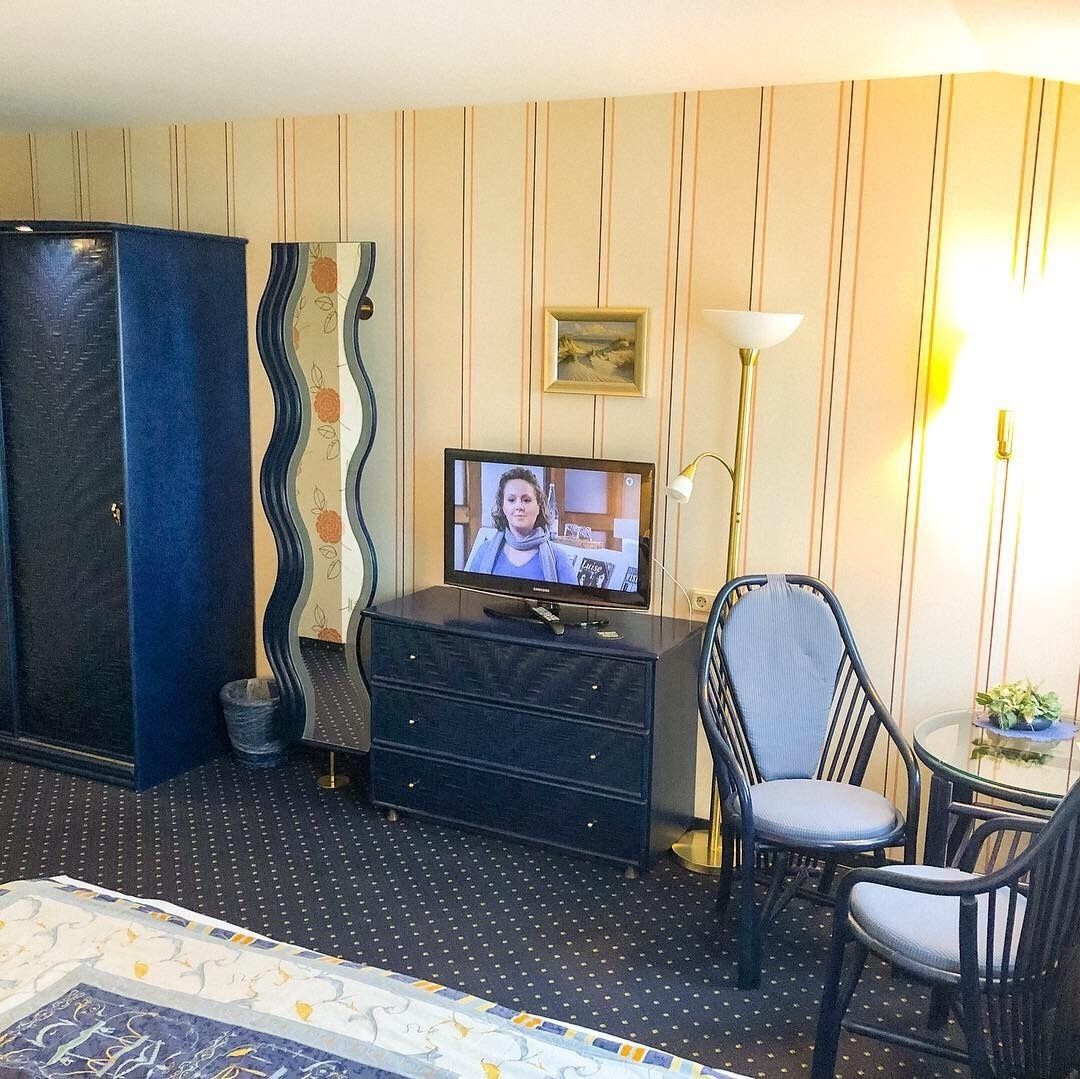 A hotel room with a television on a dresser, a chair, a small table with a lamp, and a patterned rug.