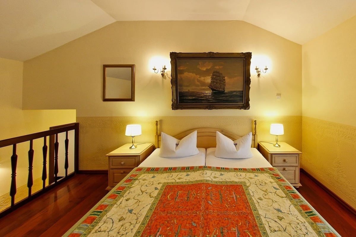A hotel room with a large bed, two bedside tables with lamps, a painting on the wall, and a view through a window.