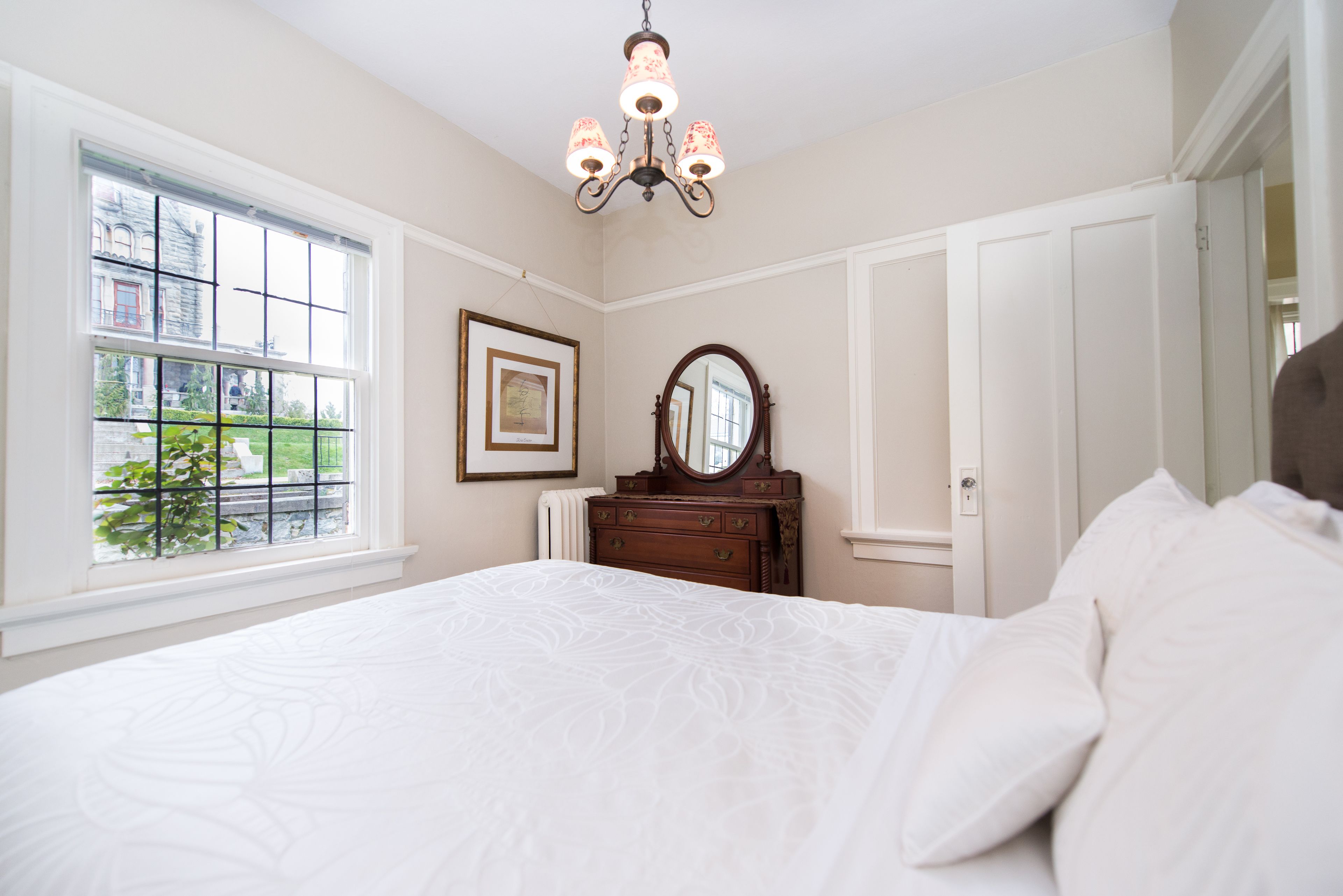 Une chambre à coucher avec un grand lit, une commode avec un miroir, un lustre et une fenêtre donnant sur un paysage verdoyant.