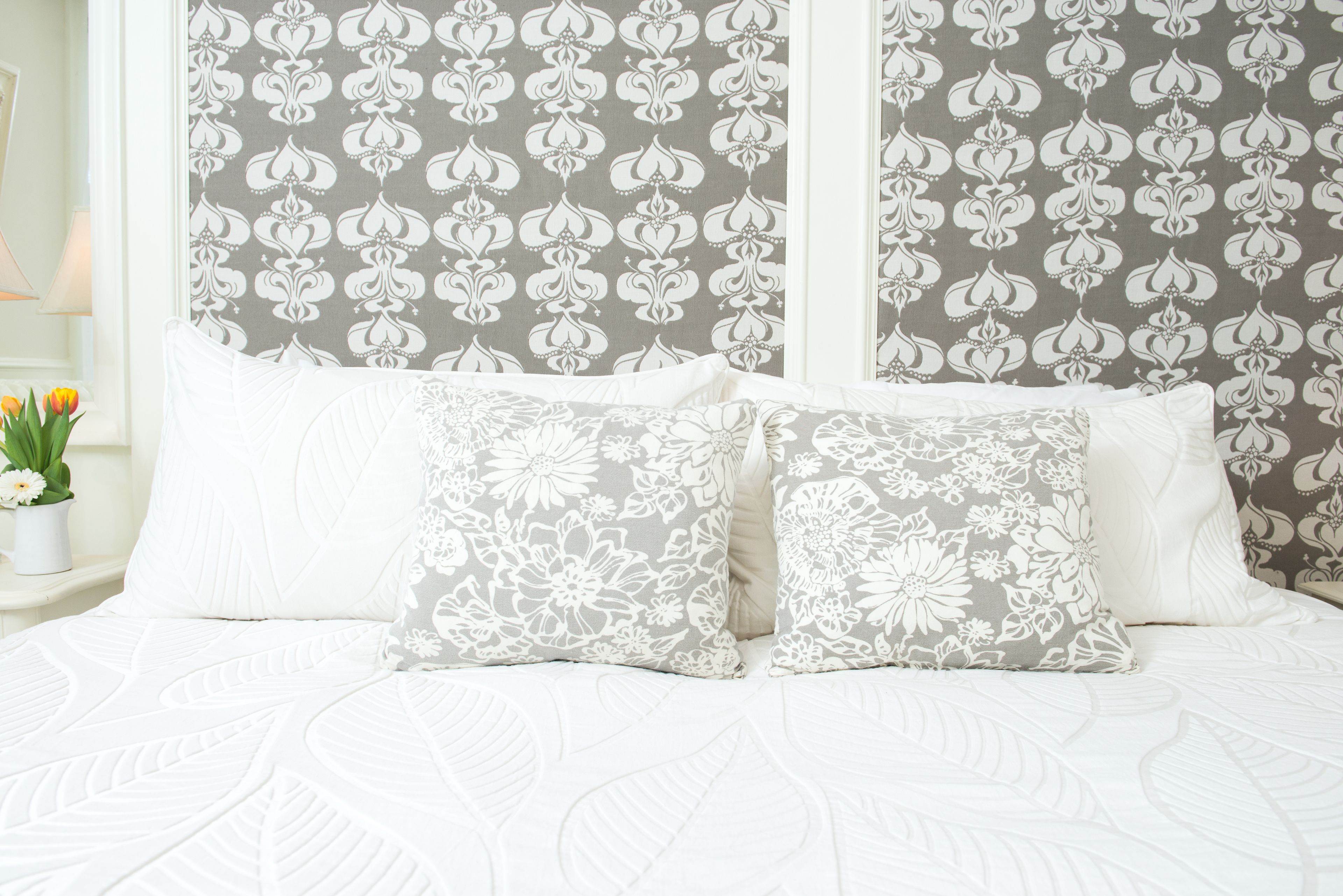 Una cama con ropa blanca y almohadas estampadas con motivos florales, frente a una pared con un papel tapiz de diseño floral repetido.