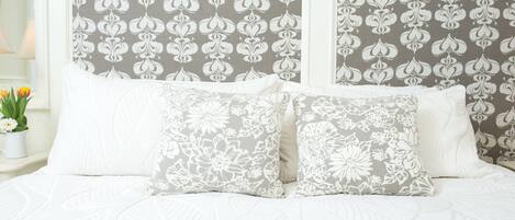A bed with white bedding and floral-patterned pillows, set against a wall with a repeating floral wallpaper pattern.