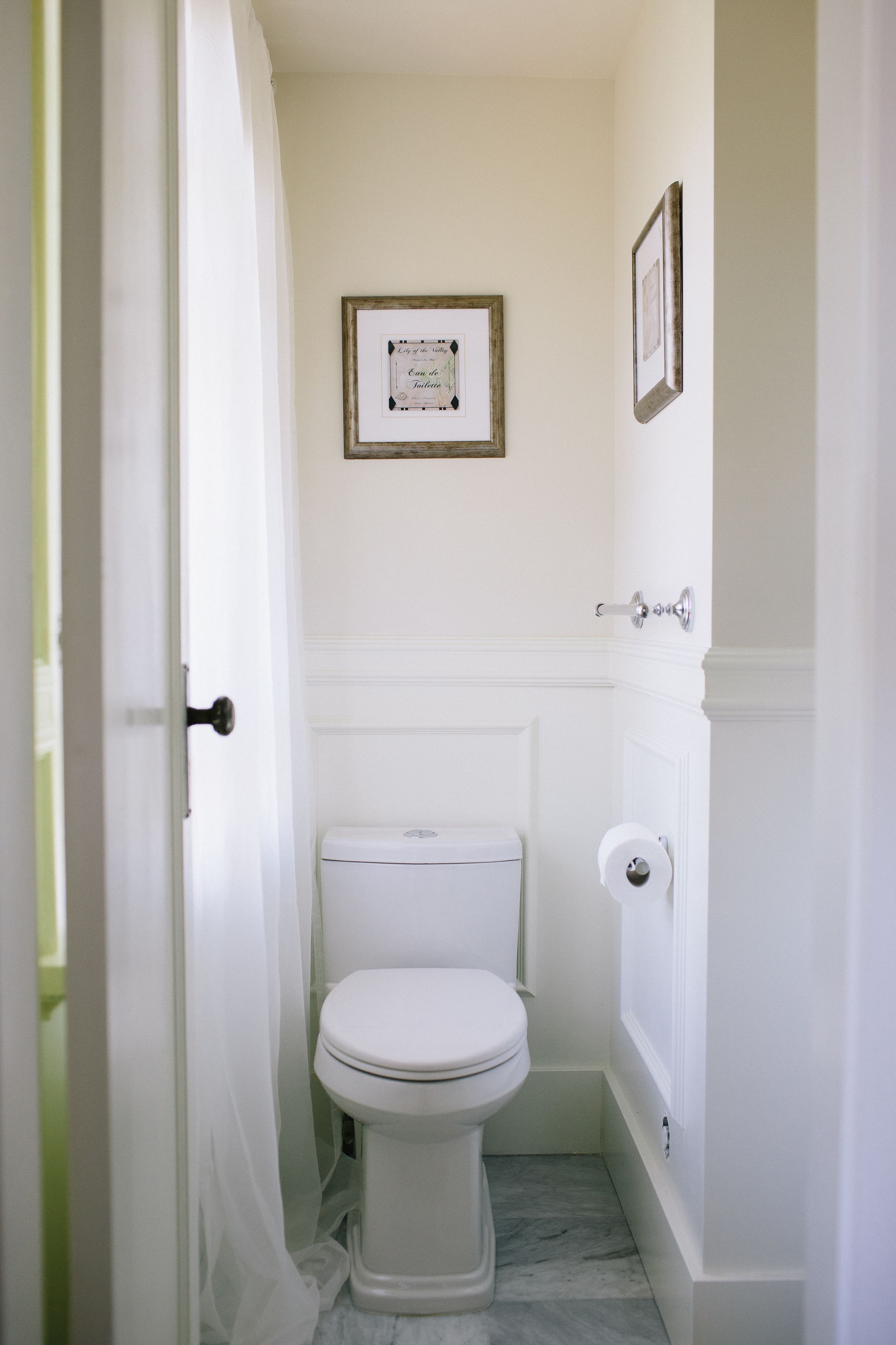 Une petite salle de bain blanche, avec des toilettes, un certificat encadré accroché au mur et une porte munie d’un rideau.