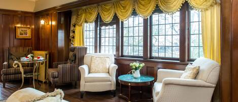 A room with wooden paneled walls, a large window with yellow curtains, and a seating area with two armchairs and a small table.