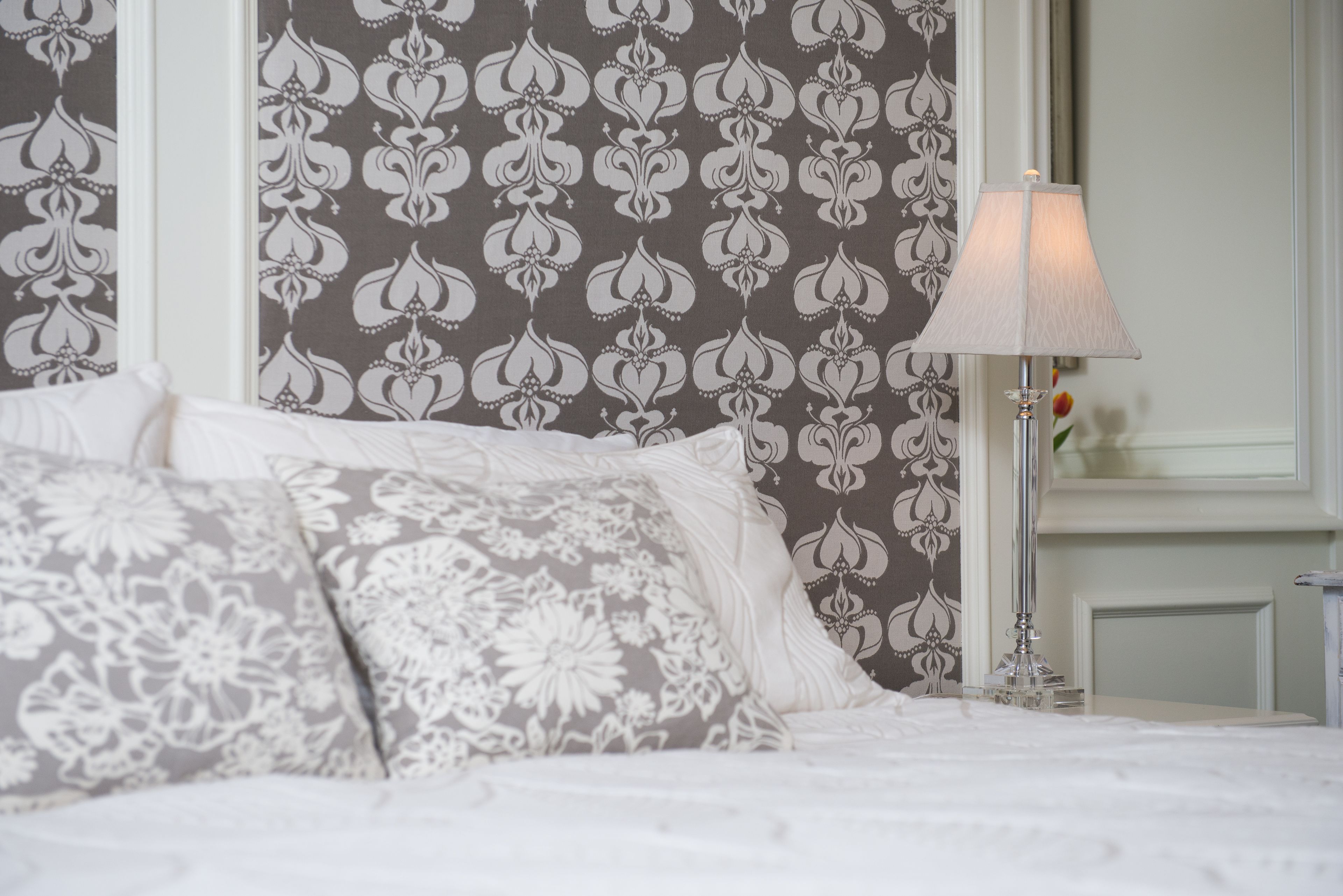 A neatly made bed with patterned pillows, a bedside lamp, and a wall with a floral wallpaper design.