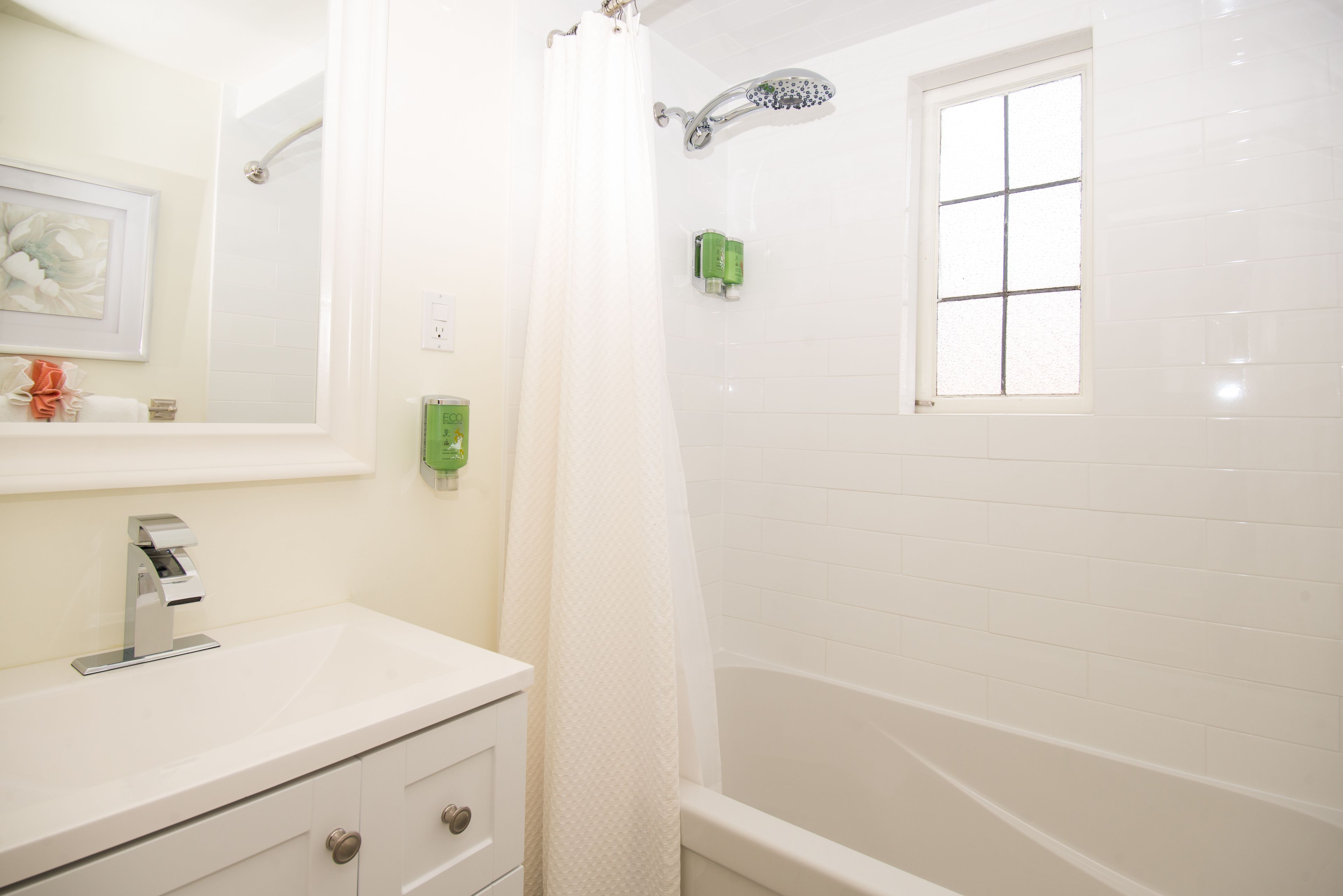 Un baño con lavamanos blanco, espejo, ducha con cortina y ventana.