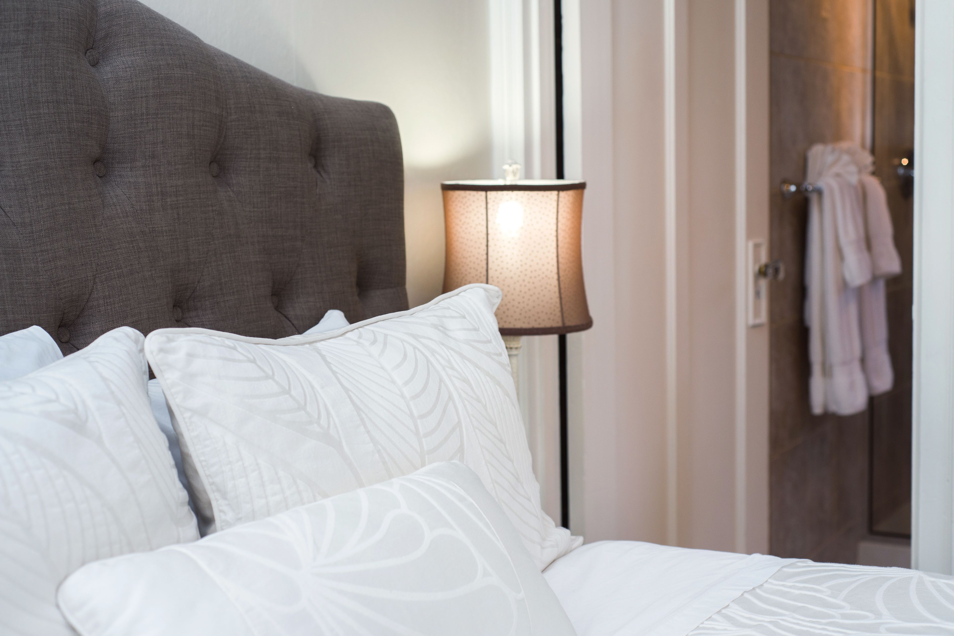 A neatly made bed with white linens and a tufted headboard, a bedside lamp, and a door with a towel hanging on it.
