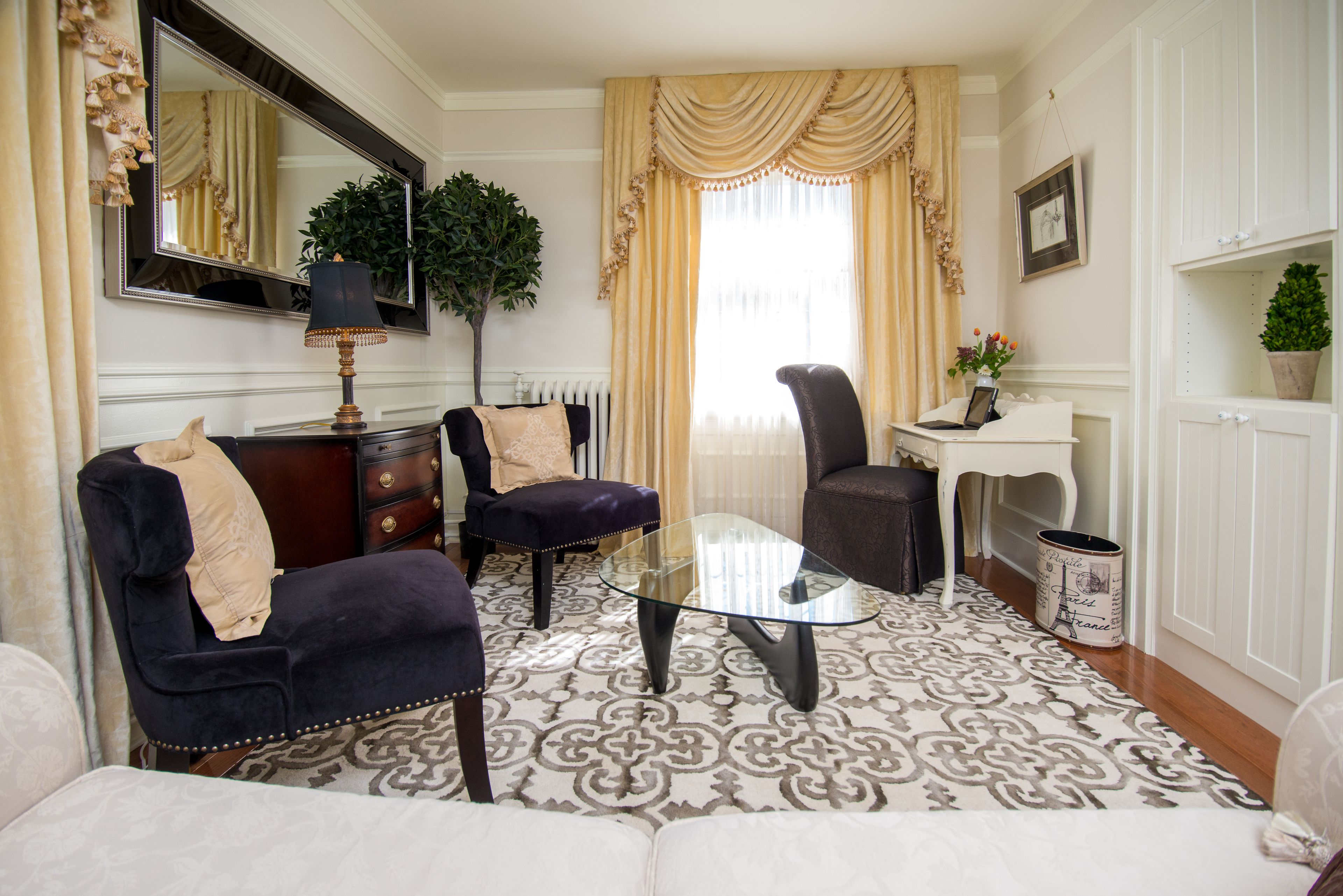 Un salon comprenant une table basse en verre, deux fauteuils, un canapé, une commode, un miroir, une plante en pot et un radiateur.