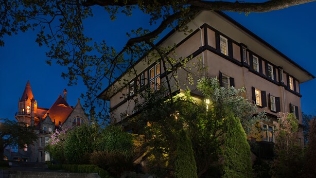 Un edificio grande e histórico con varios pisos y una torre destacada, rodeado de vegetación exuberante y jardines bien cuidados.