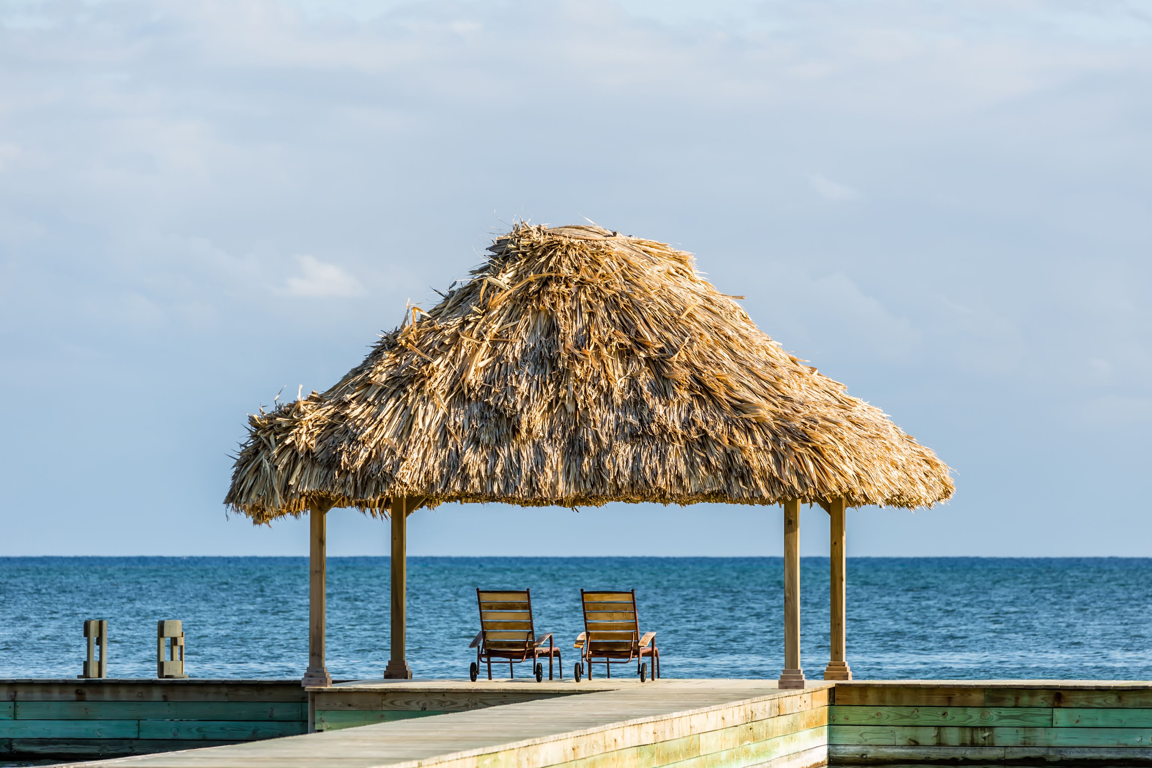 A wooden deck with two lounge chairs under a thatched umbrella, overlooking the sea.