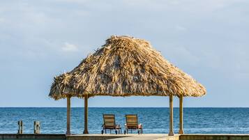 A wooden deck with two lounge chairs under a thatched umbrella, overlooking the sea.