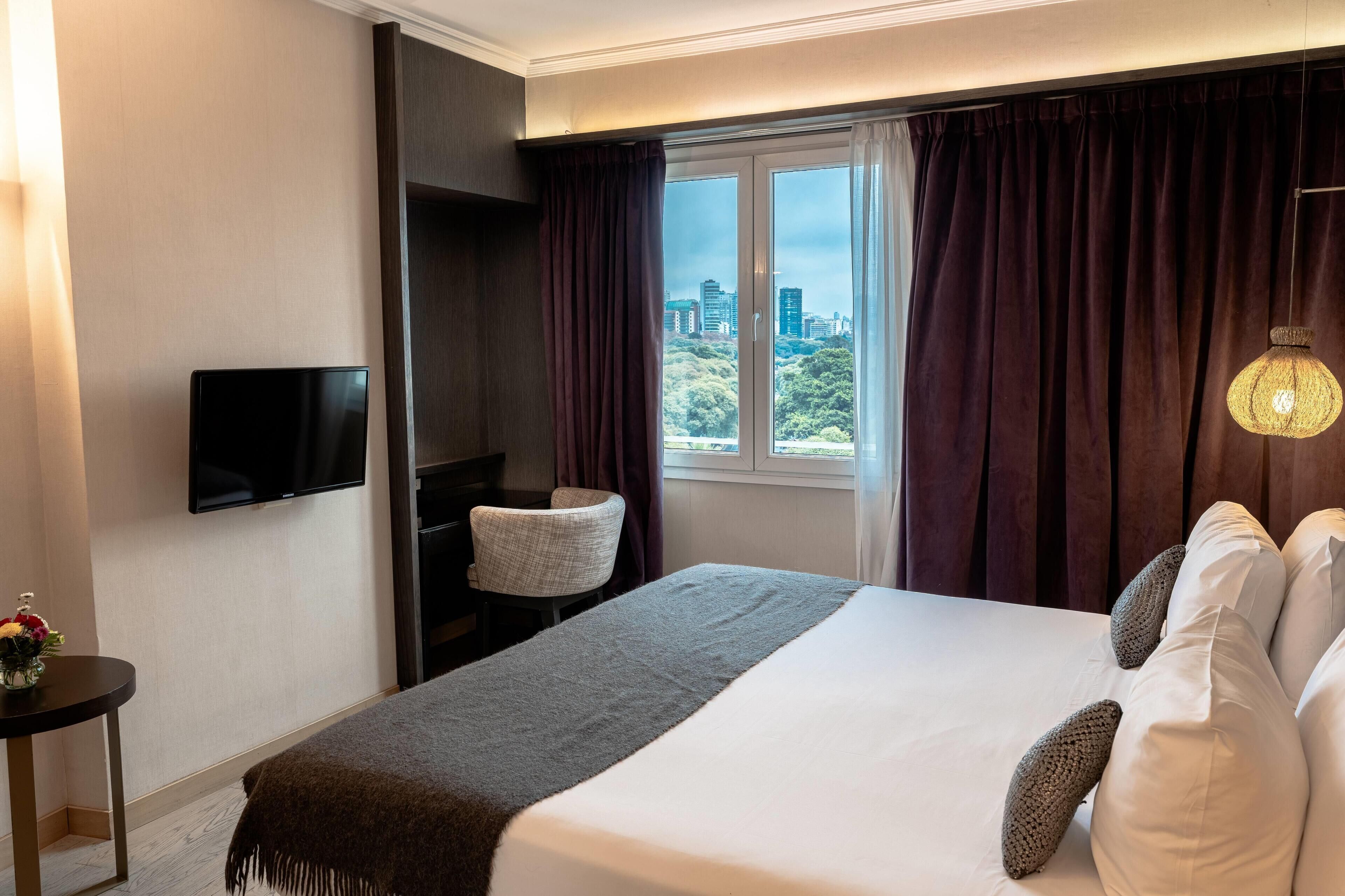 A hotel room with a bed, a TV, a chair, a window with curtains, and a view of a cityscape.