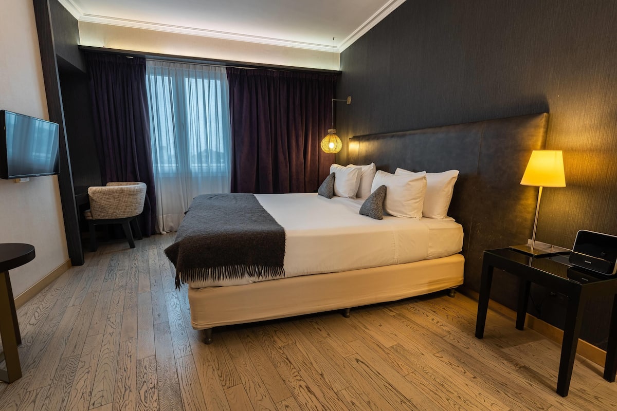 Ein Hotelzimmer mit einem großen Bett, einem Nachttisch mit Lampe, einem Sessel, einem Fernseher und Blick ins Freie durch Vorhänge.