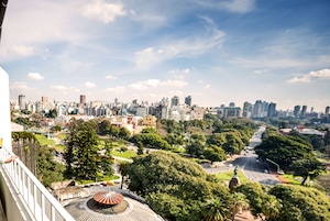 Un paisaje urbano que combina edificios modernos y antiguos, un parque con árboles y una estatua, y un cielo despejado.