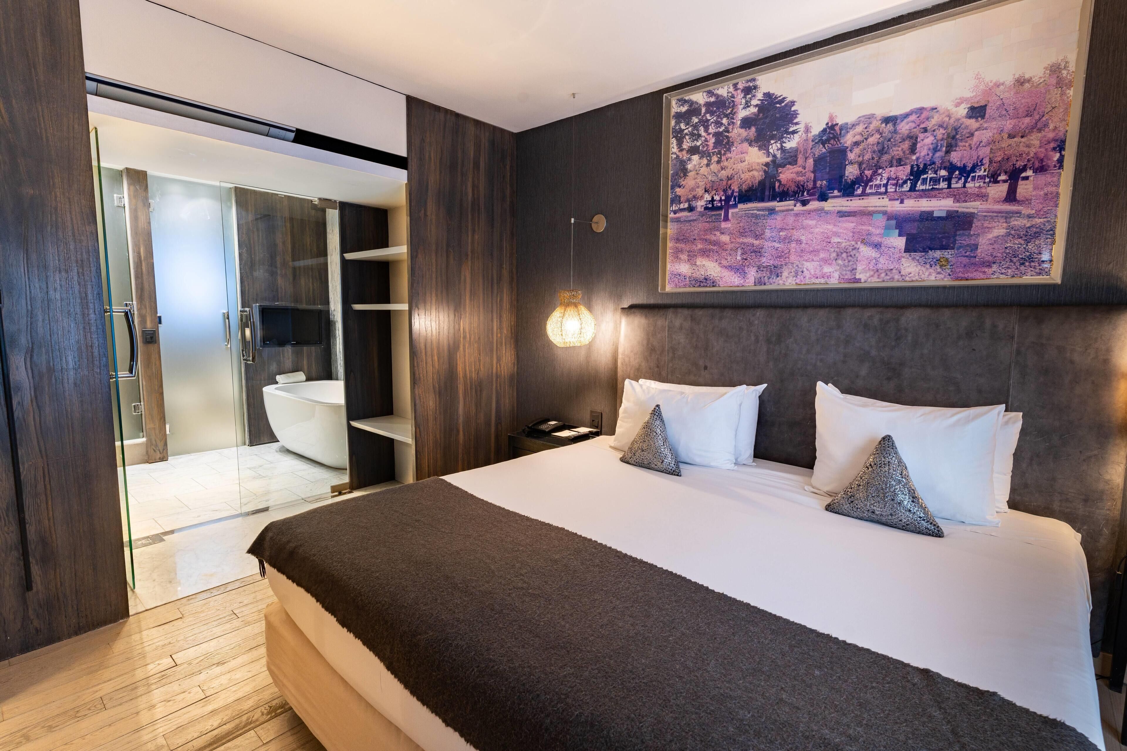 Una habitación de hotel moderna con una cama grande, un cuadro de un parque y una zona de ducha con paredes de vidrio.