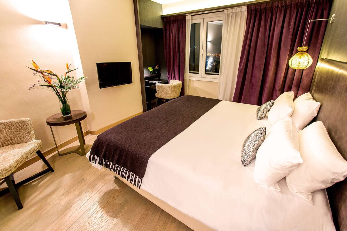 Ein Hotelzimmer mit einem großen Bett, einem Sessel, einem Schreibtisch, einem Fernseher und einem Fenster mit Vorhängen.