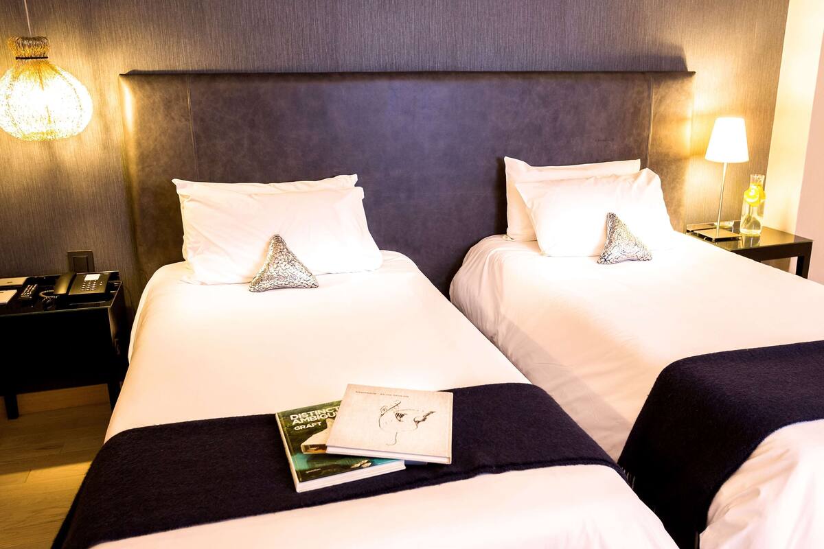 Ein Hotelzimmer mit zwei Betten, jeweils mit weißer Bettwäsche und dekorativen Kissen. Am rechten Bett befindet sich ein Nachttisch mit Telefon und Lampe. Auf der linken Seite des Bettes liegt ein Buch.