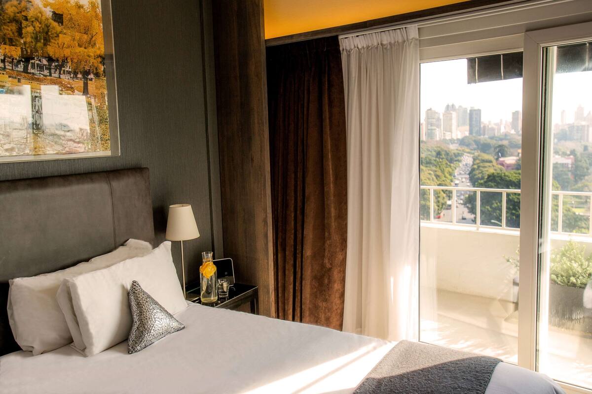 Ein Hotelzimmer mit einem Bett, einem Nachttisch mit Lampe, einem Fenster mit Vorhängen und Blick auf die Stadtlandschaft.