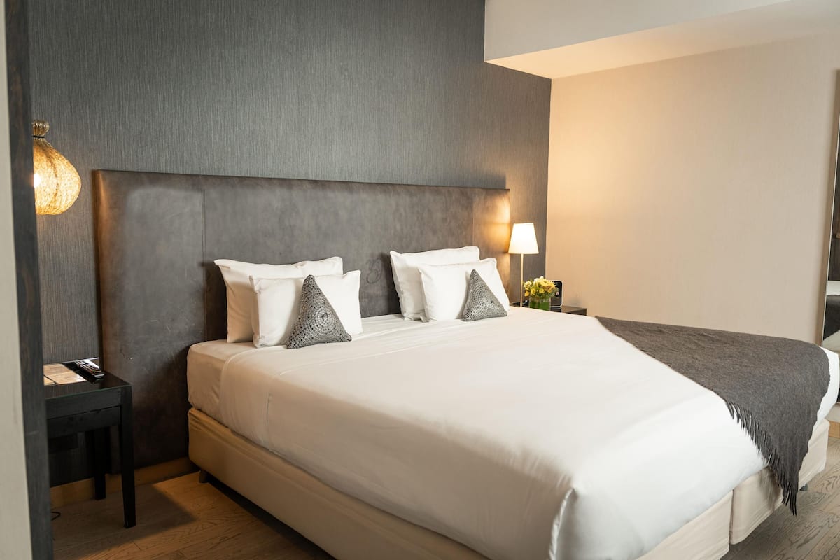 Ein modernes Hotelzimmer mit einem großen Bett, einem dunklen Kopfteil, einem Nachttisch mit einer Lampe und einem kleinen Tisch.