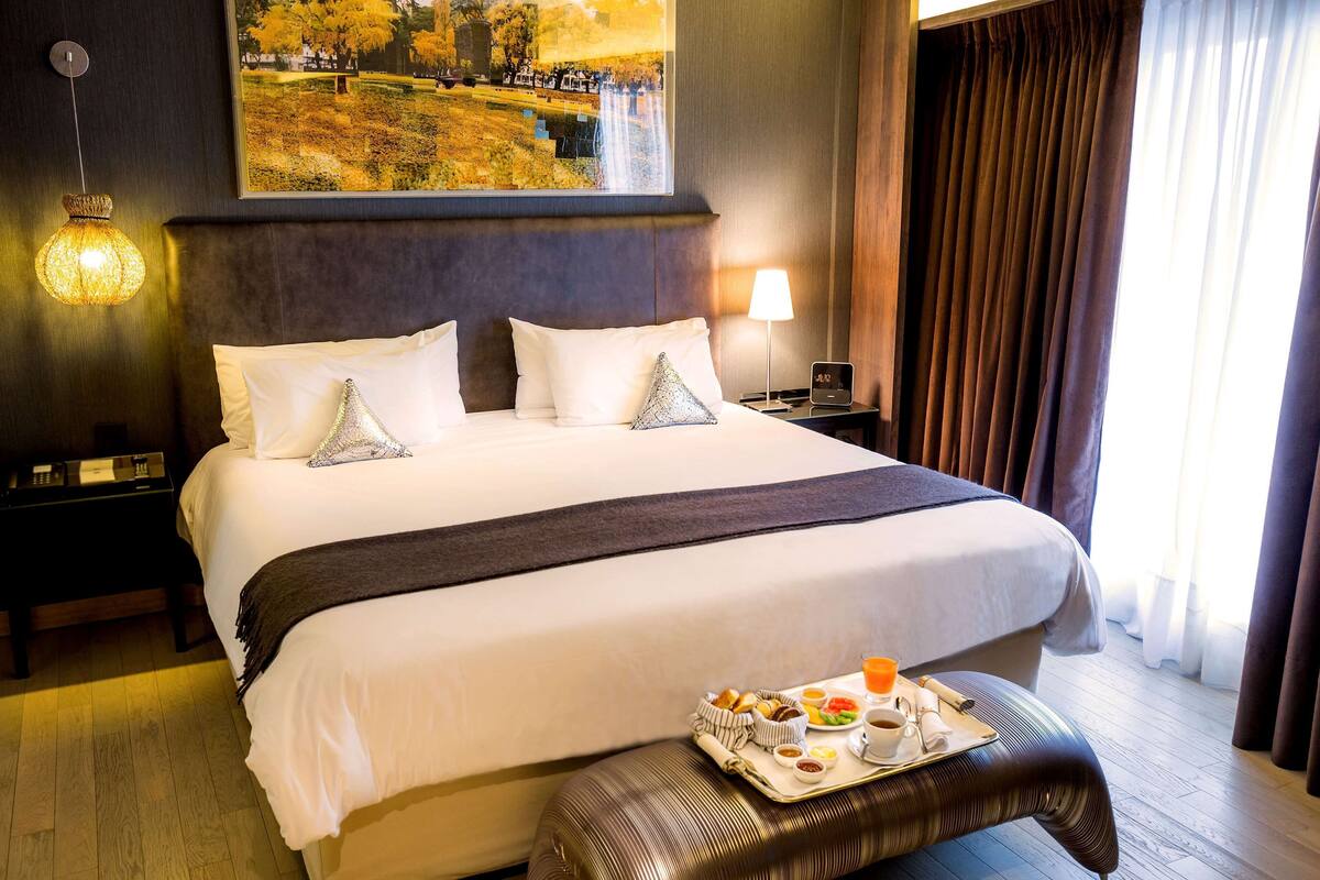 Ein Hotelzimmer mit einem großen Bett, einem Nachttisch mit Lampe, einem Tablett mit Frühstücksgebäck und einem Bild an der Wand.