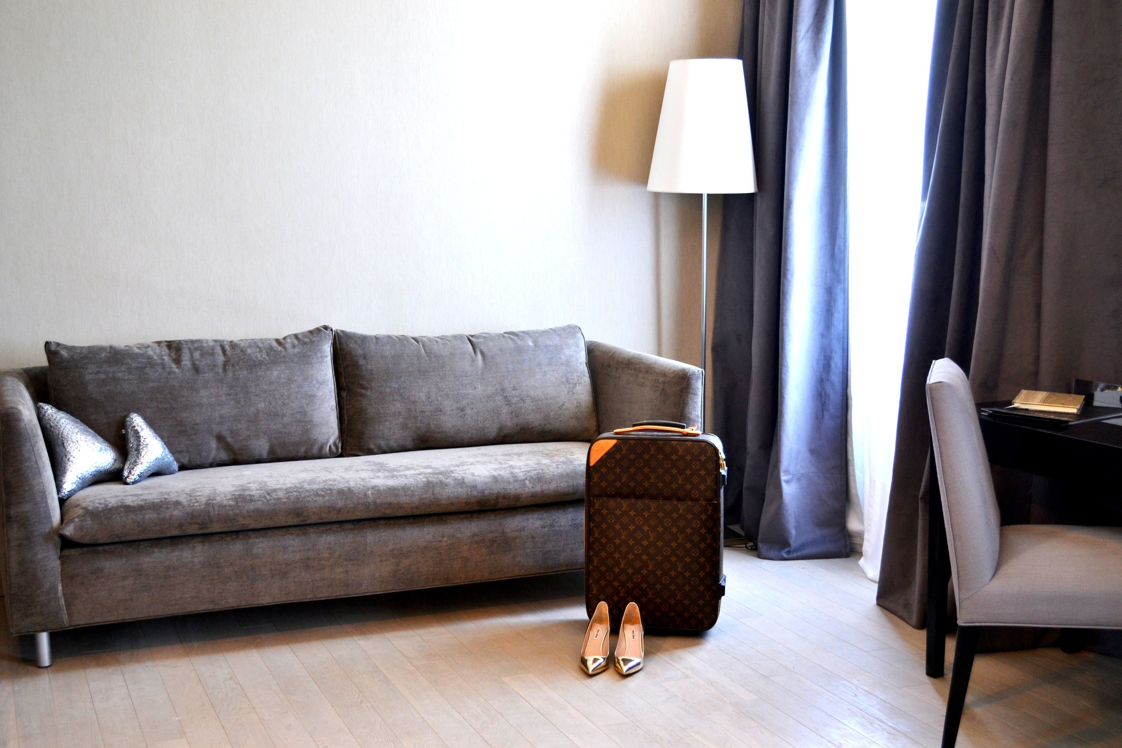A grey sofa with two pillows, a brown suitcase, a pair of shoes, a lamp, and a chair in a room with wooden flooring.