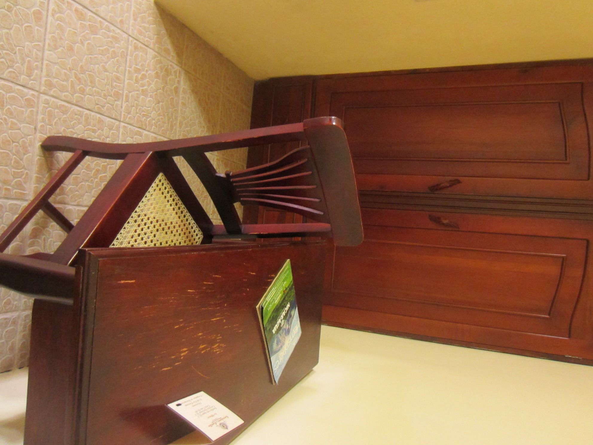 A wooden chair with a backrest and armrests, placed against a wall with a patterned tile design.