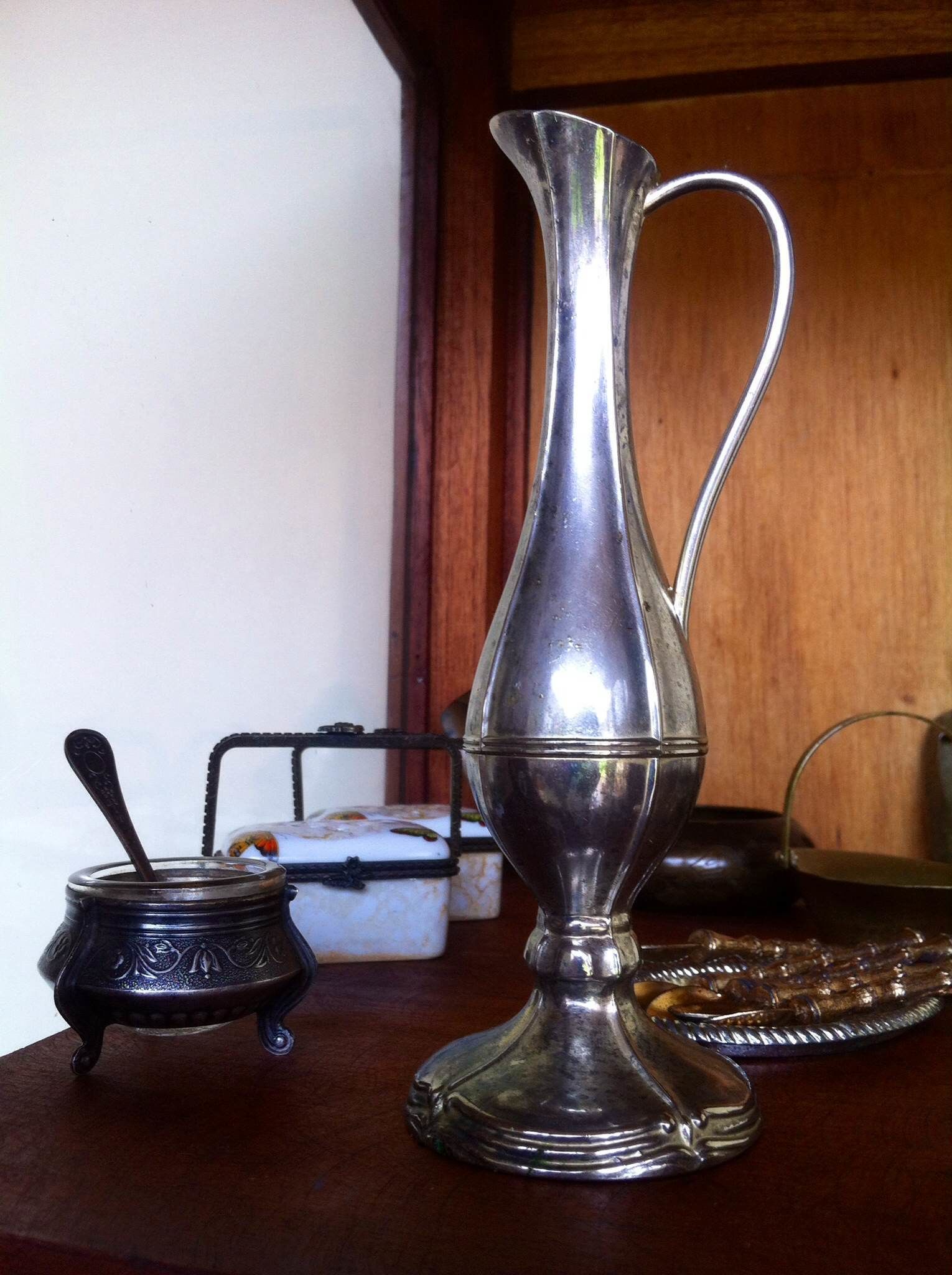A silver pitcher with a handle, a small metal container with a spoon, and a glass on a wooden surface.