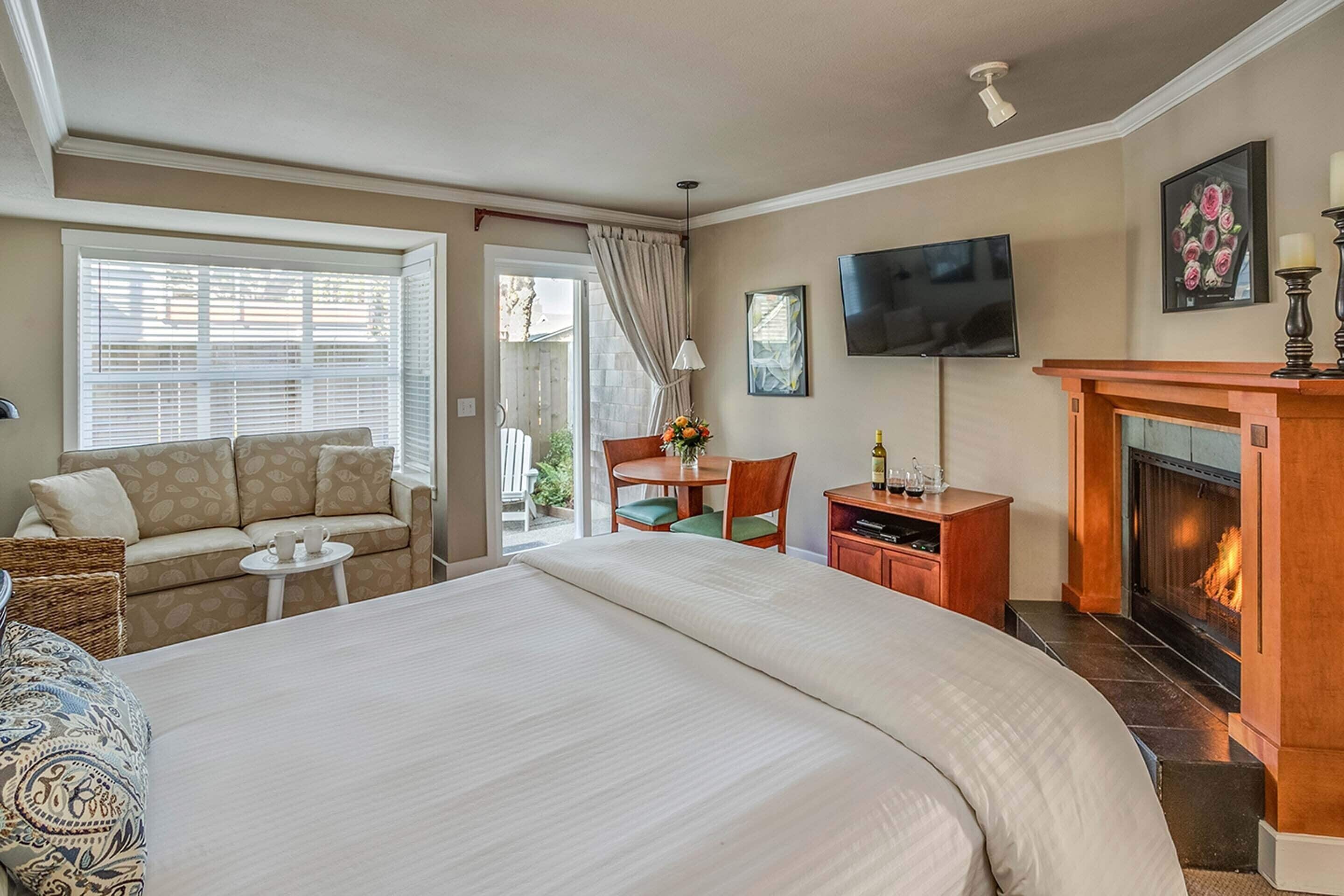 Habitación de hotel con una cama grande, un sofá, una mesita con sillas, un televisor, una chimenea y una ventana con persianas.