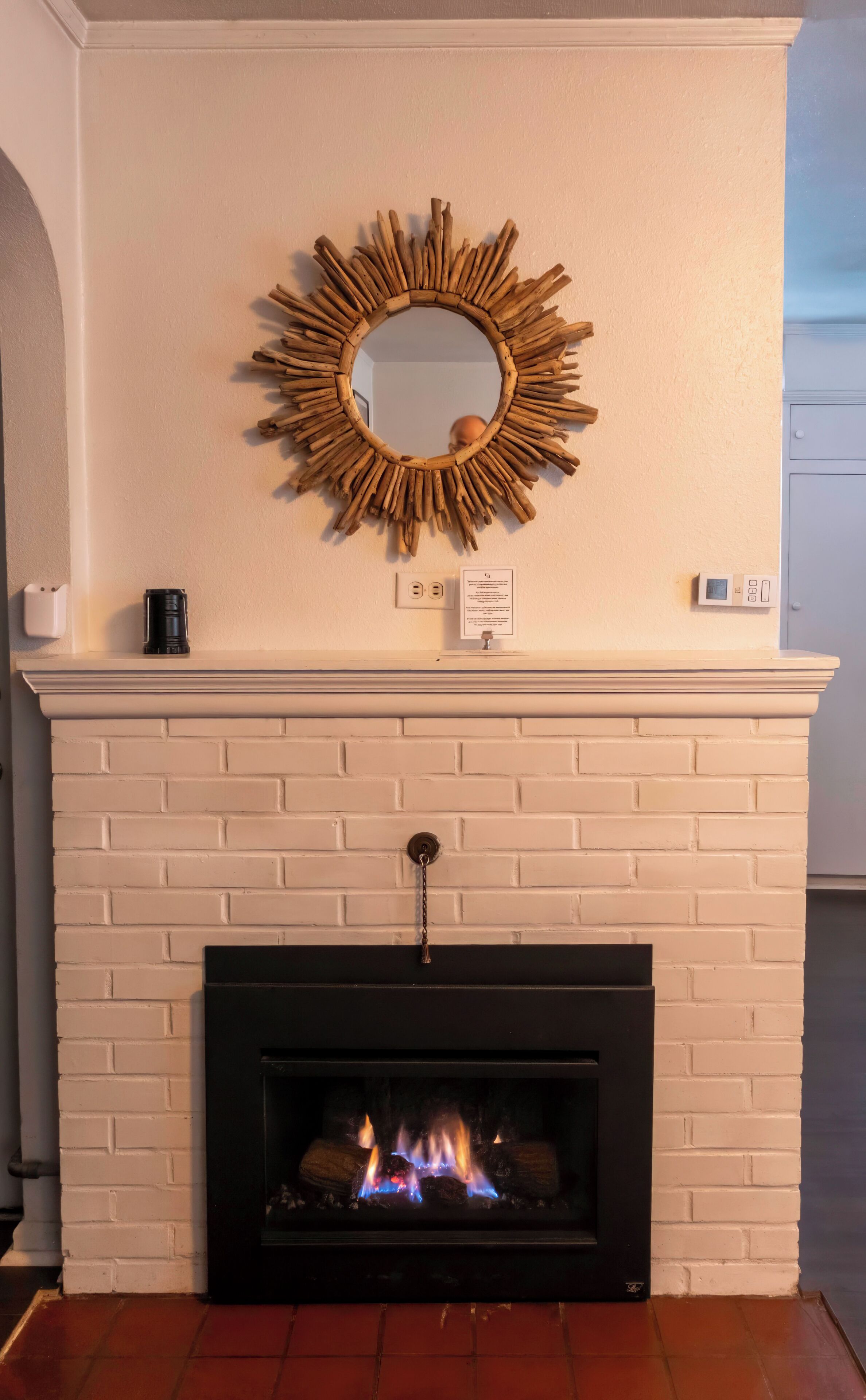 A fireplace with a brick mantel, a mirror, and a thermostat on the wall.