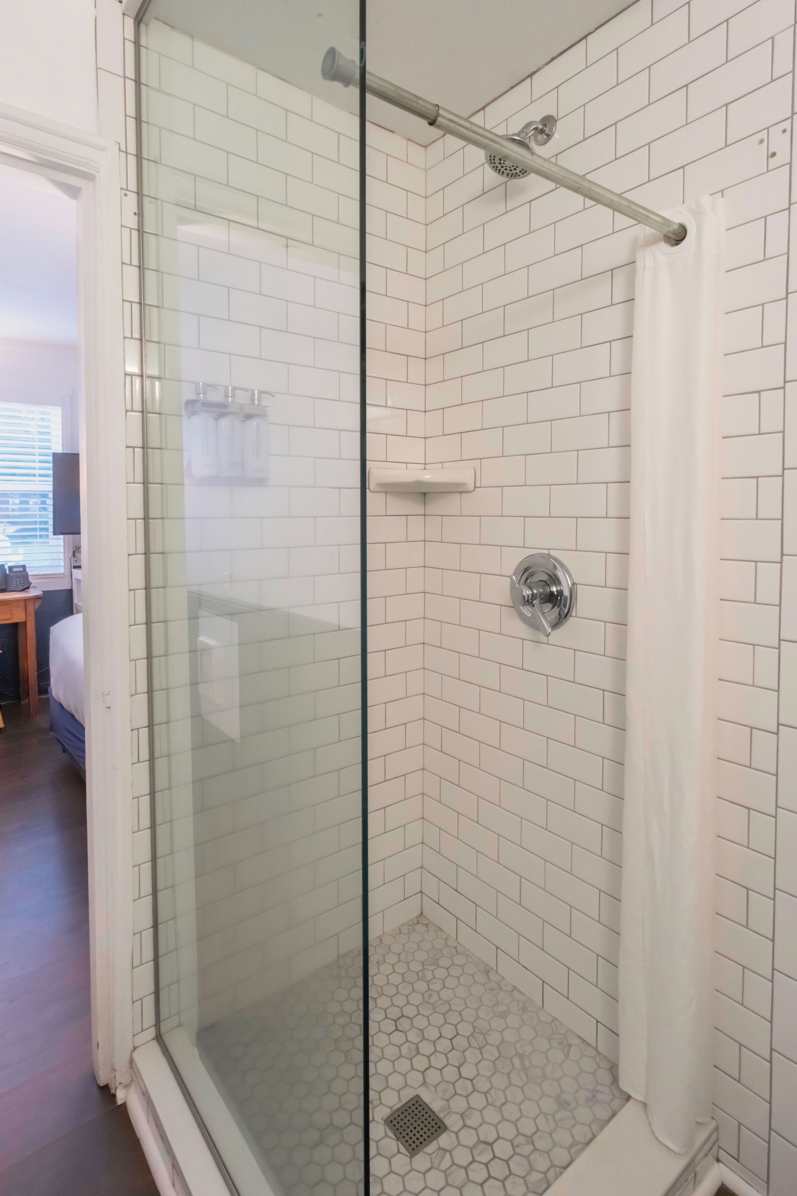 Una ducha con cerramiento de vidrio, paredes de azulejos blancos y un patrón de suelo hexagonal.