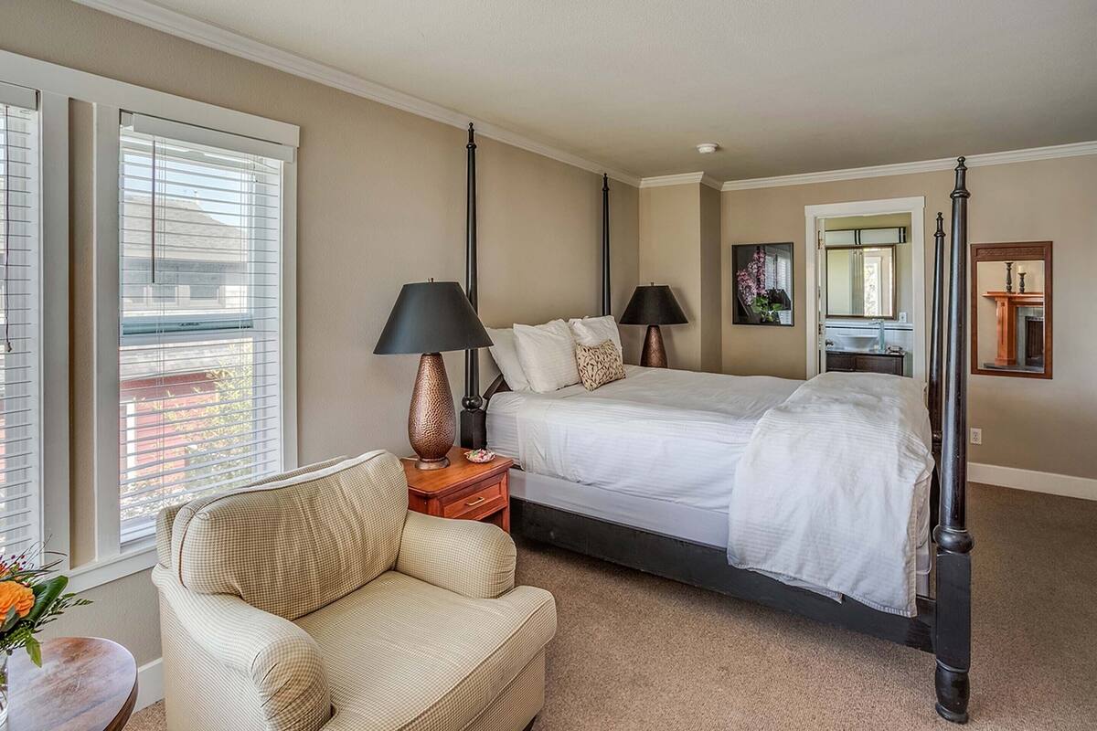 A bedroom with a four-poster bed, a bedside table with lamps, a chair, and a window with blinds.
