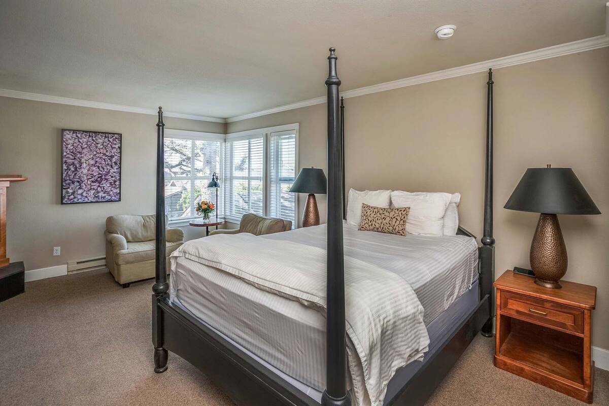 A four-poster bed with a wooden frame, a beige carpet, a window with blinds, a chair, and a lamp on a wooden nightstand.
