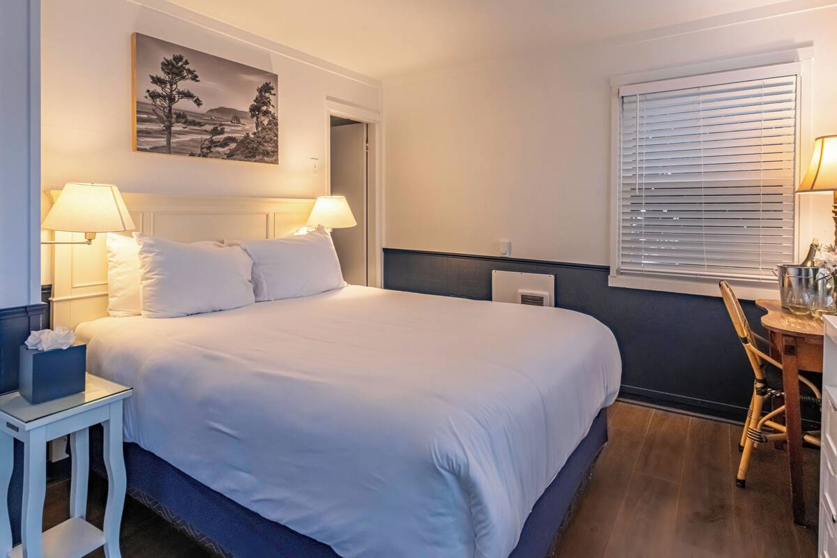 A hotel room with a bed, two bedside lamps, a bedside table with a tissue box, a chair, a window with blinds, and a painting on the wall.