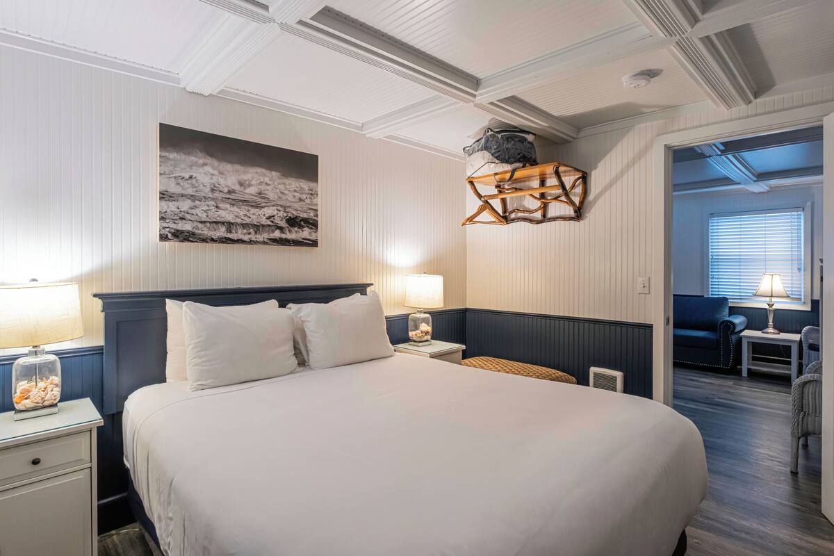 A hotel room with a large bed, two bedside tables, a wall-mounted lamp, a decorative wall piece, and a view into the adjacent living area.
