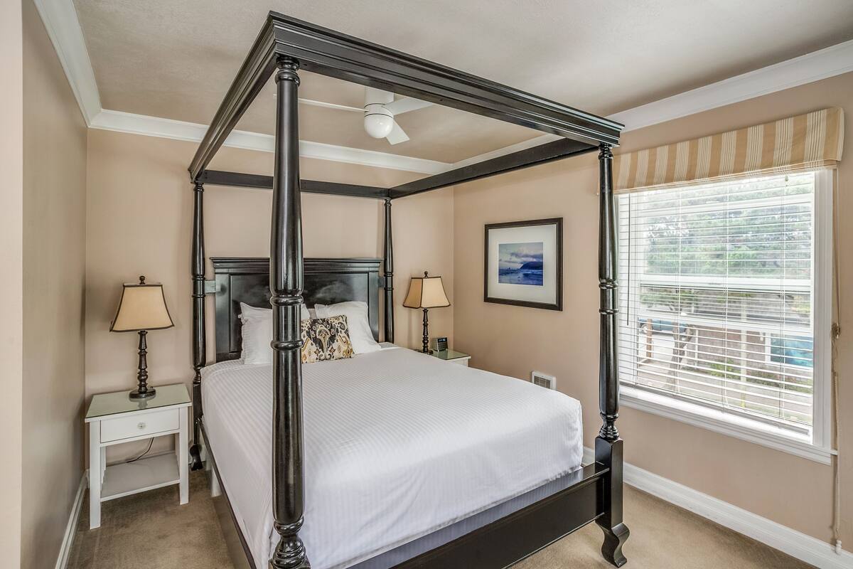 A four-poster bed with a headboard, two bedside tables, a nightstand, a ceiling fan, a window with blinds, and a framed picture on the wall.