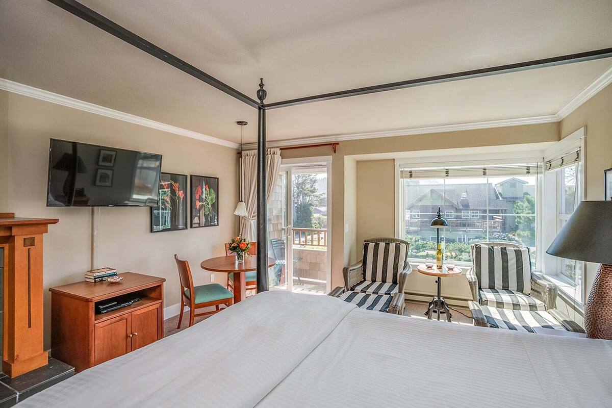 A hotel room with a bed, a desk, a chair, a small table, and a balcony with a view.