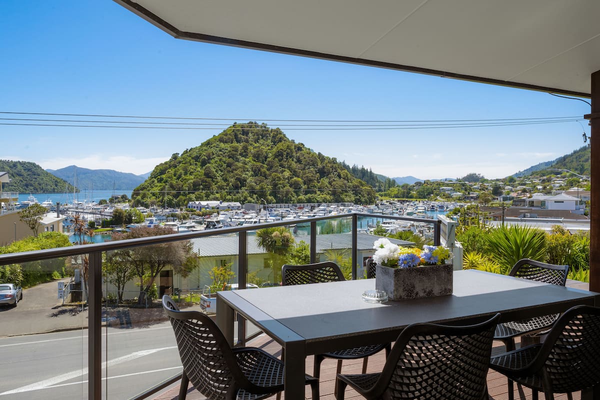 A balcony with a table and chairs, overlooking a marina with boats and a hill with greenery.