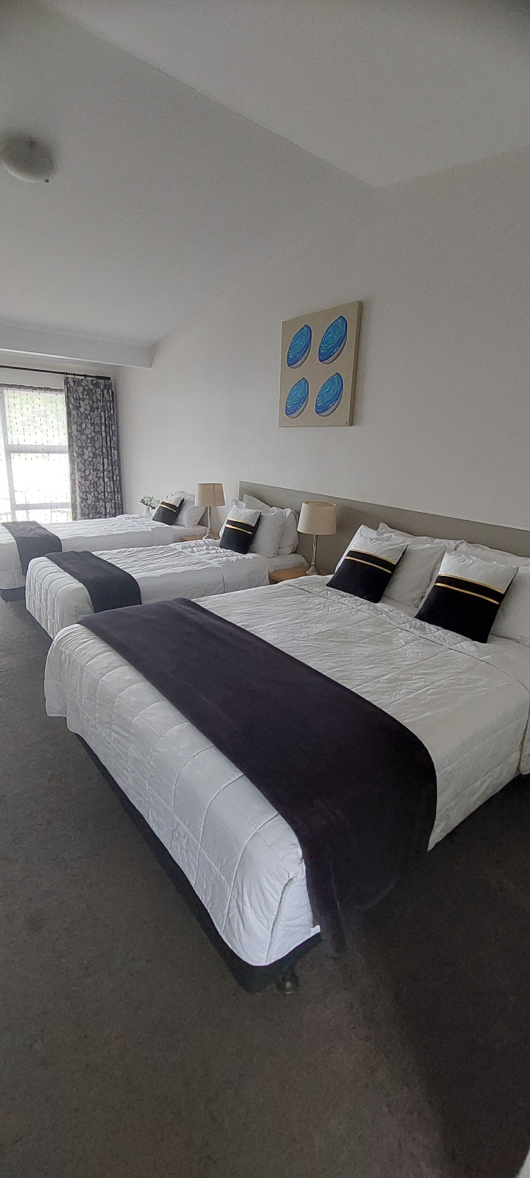 A hotel room with three beds, a large window with curtains, a painting on the wall, and a lamp on the nightstand.