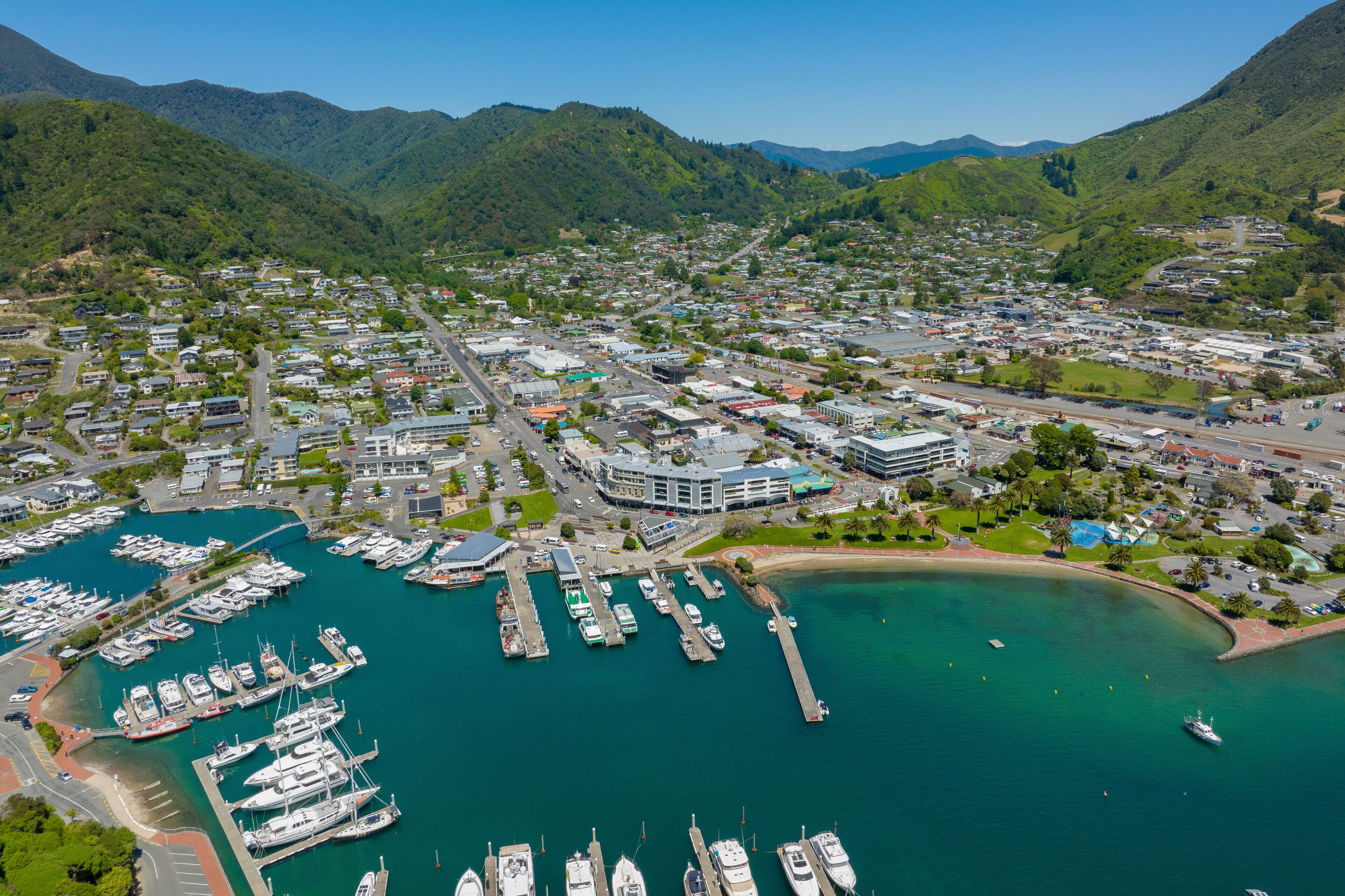 An aerial view of a coastal town with a marina, buildings, and mountainous backdrop.