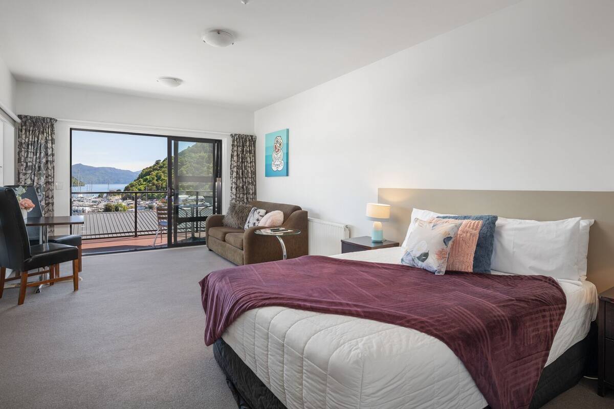 A hotel room with a bed, a desk, a chair, a nightstand, a lamp, and a view of the outdoors.