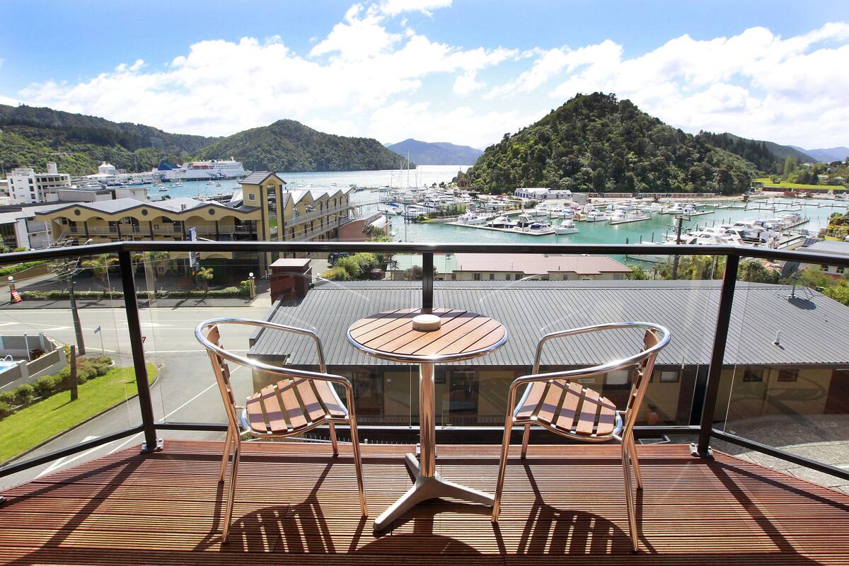 A balcony with two chairs and a table, overlooking a marina with boats and a hilly landscape.