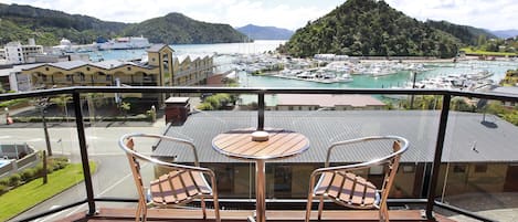 A balcony with two chairs and a table, overlooking a marina with boats and a hilly landscape.