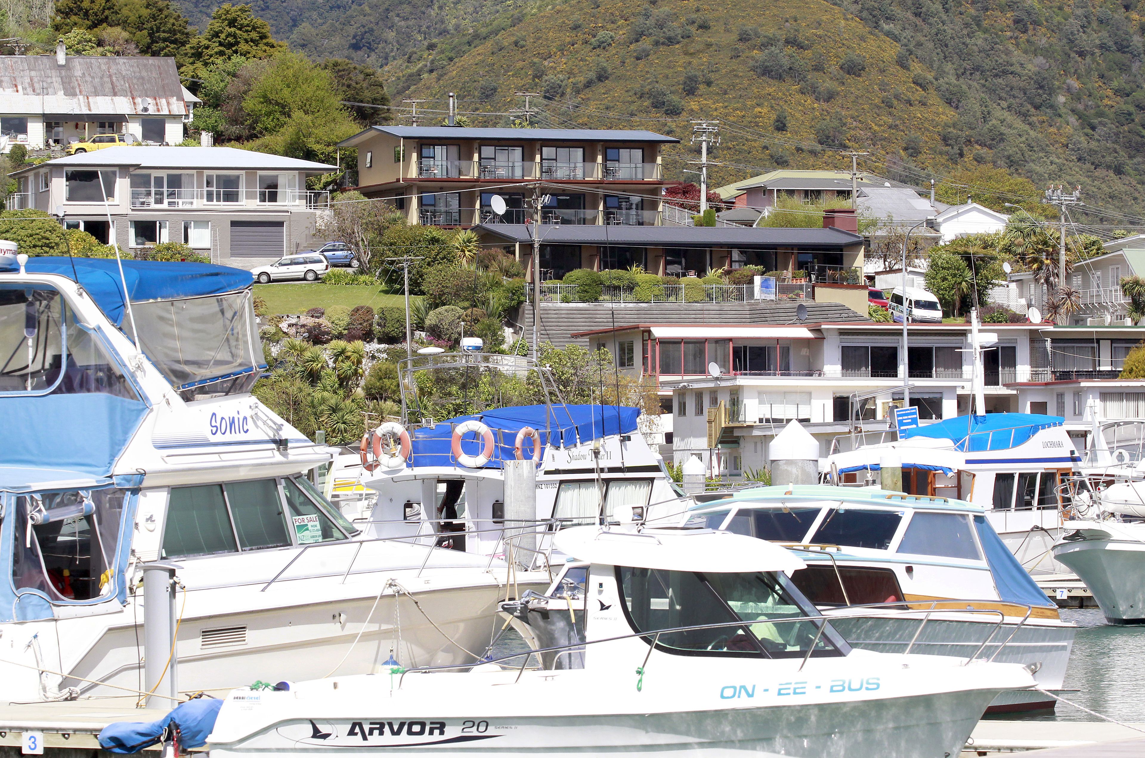 A marina with several boats docked, surrounded by residential buildings and a mountainous backdrop.