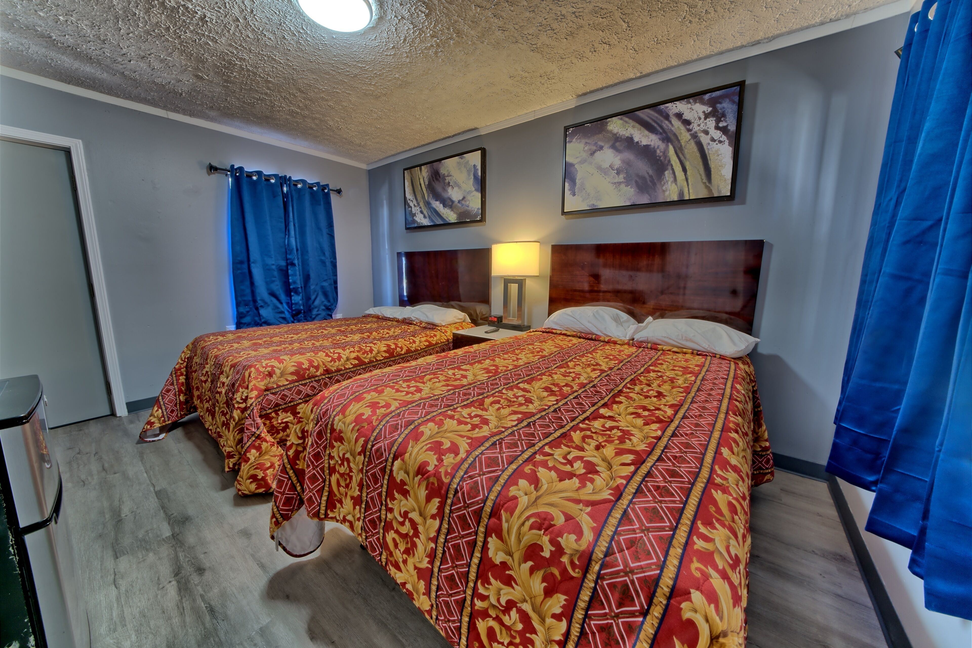 Quarto de hotel com duas camas, uma cabeceira de madeira, uma mesinha de cabeceira, uma luminária e uma janela com cortinas azuis.
