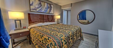 Un cuarto de hotel con una cama grande, dos mesitas de noche con lámparas, un espejo redondo y un cuadro en la pared.